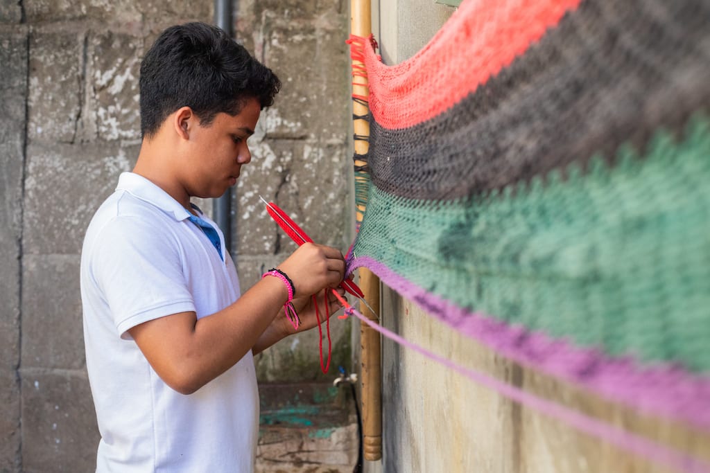 A boy in a white t-shirt woks on a red, black, green and purple hammock.